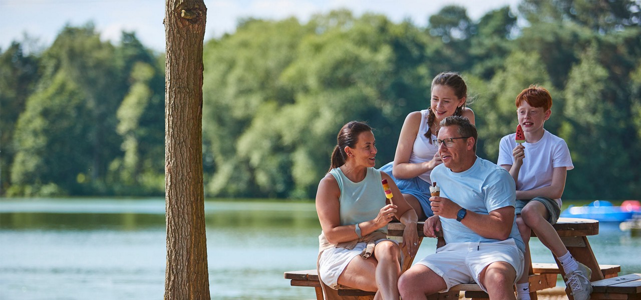 Family of four sit and eat ice lollies, chatting and smiling, on a wooden picnic bench beside a calm lake; trees, distant boats, and summer light create a relaxed setting.