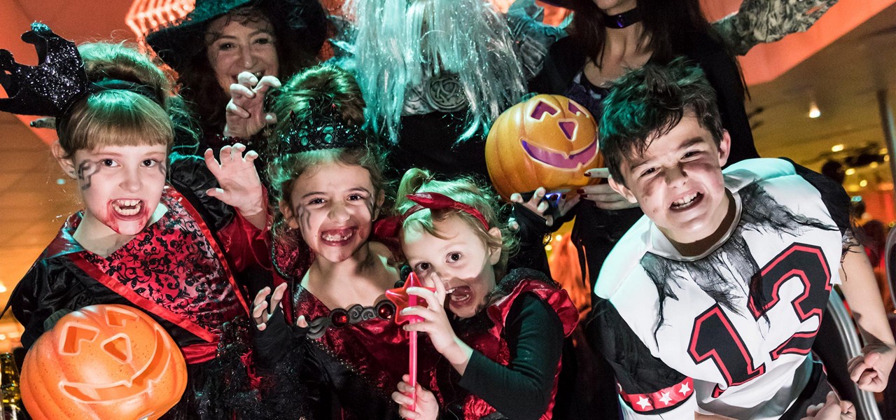 Costumed children pose and grin, showing fake fangs and makeup, while holding jack-o'-lantern buckets and wands; adults in spooky outfits stand behind indoors. Visible text: 13 on a boy’s shirt.