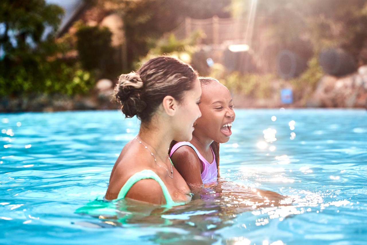 Two people—an adult and a child—laugh while wading chest-deep, facing right; sunlight sparkles on blue water, with blurred greenery and rocks around the outdoor pool.