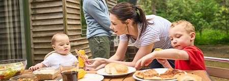 Children reach for food as their mother serves them at an outdoor picnic table; another adult stands nearby amid salad, buns, corn, pizza, and drinks in a backyard.