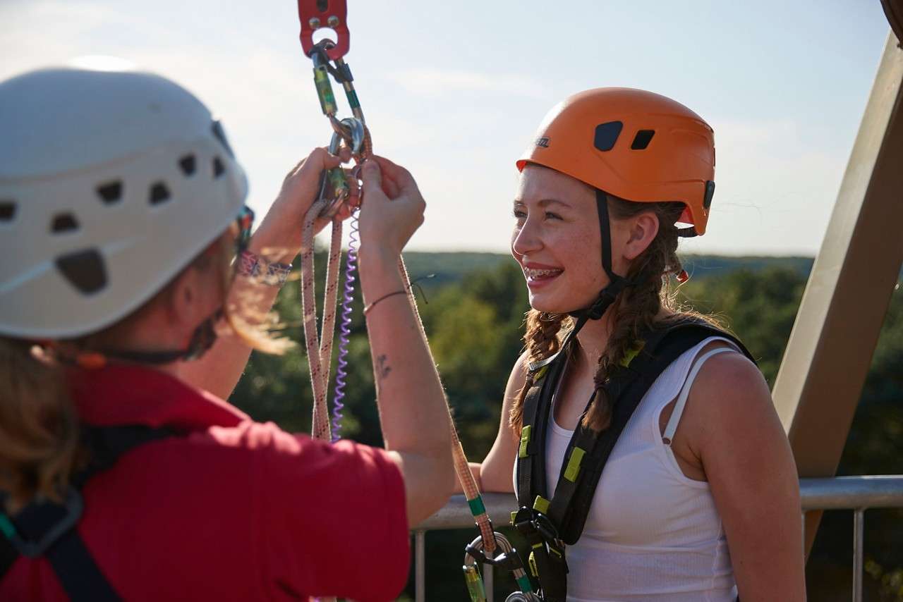Smiling teen climber waits as an instructor clips her harness to carabiners and ropes, on a high-ropes platform outdoors, with helmets and safety gear, trees and sky in the background.