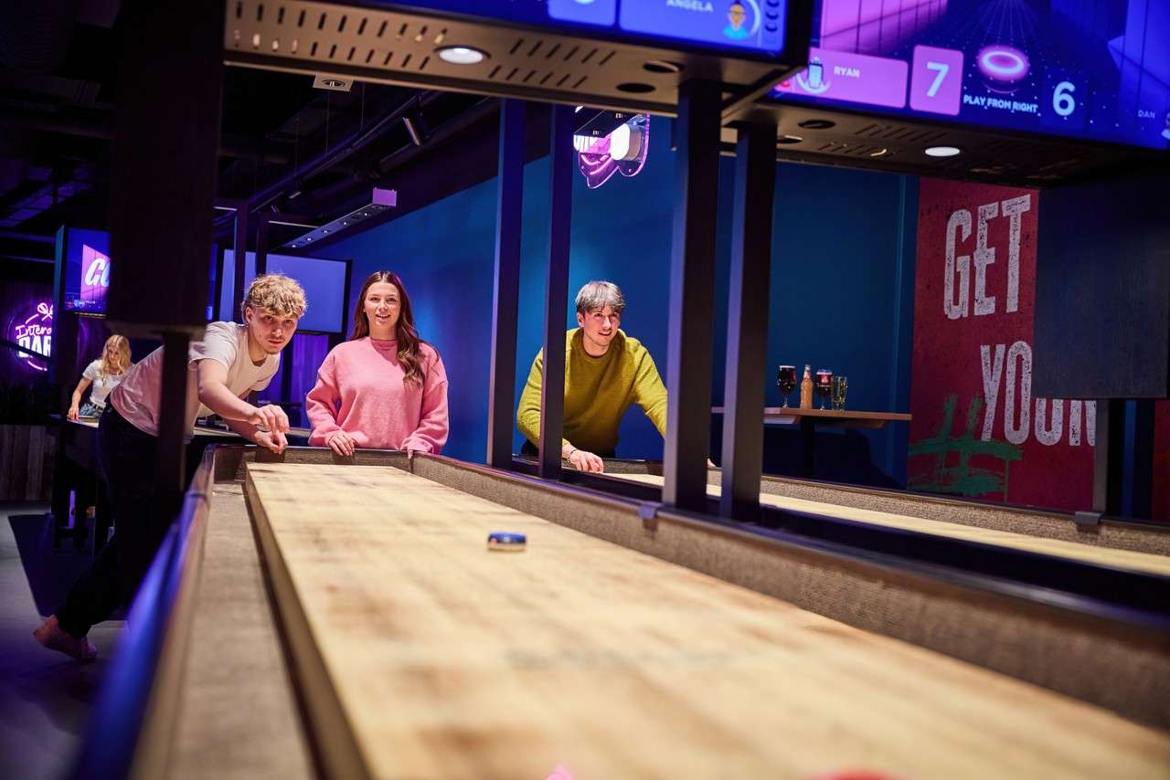 Three young adults play shuffleboard, sliding pucks toward the far end in a neon-lit game bar. Visible text: RYAN 7, DAN 6, PLAY FROM RIGHT, ANGELA, GET YOU.
