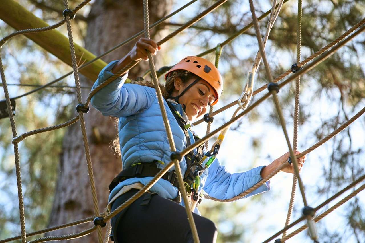 Two harnessed riders zip along parallel lines, legs splayed and arms out for balance. They traverse above a forested area at an outdoor adventure park under an overcast sky.