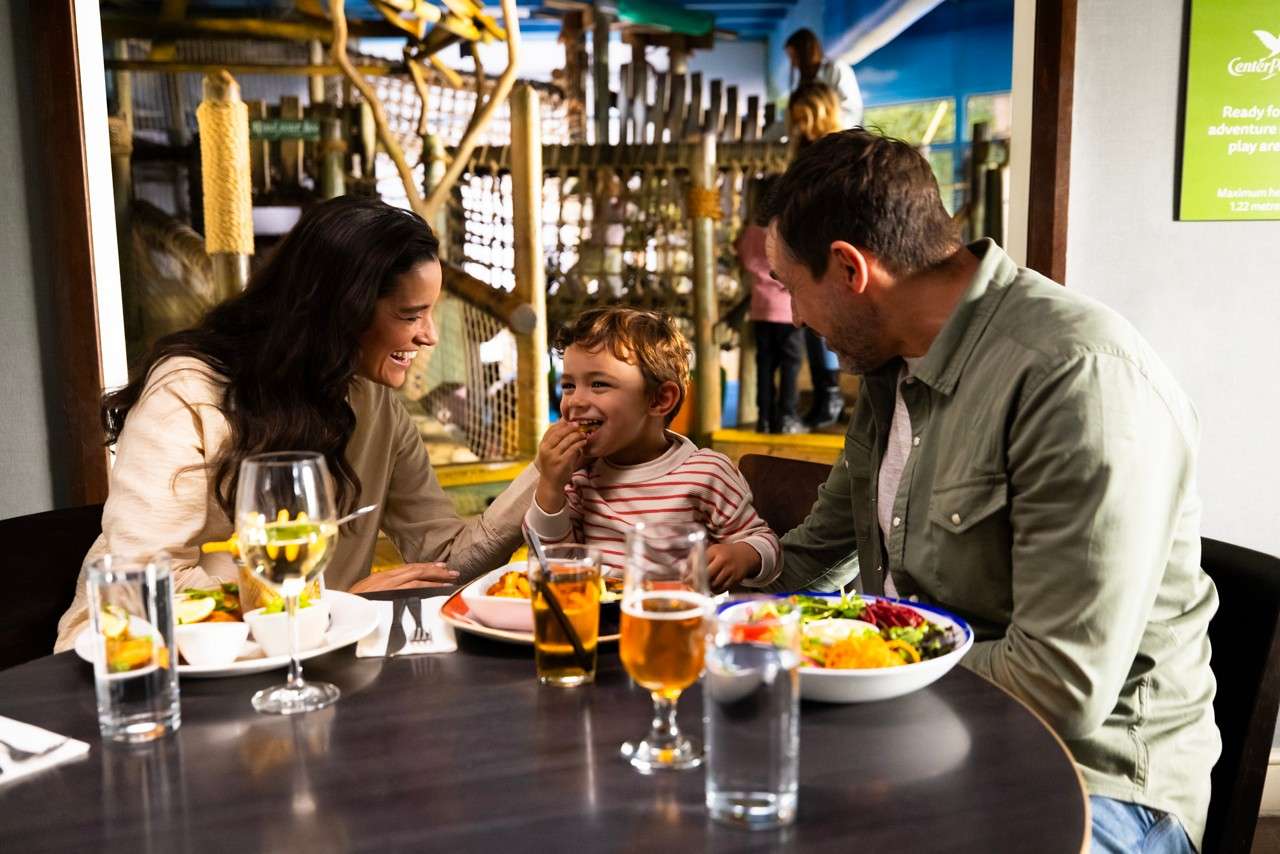 Child laughs while eating, seated between two adults at a table with salads and drinks; behind them is an indoor play area. Text on green sign: “Centre Parcs … Ready for … adventure … play area … Maximum … height 1.22 metres.”
