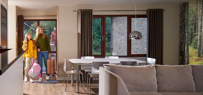 Mother and daughter enter carrying bags and a rolling suitcase; a man stands outside the door. Context: modern living-dining room with sofa, table, chairs, pendant light, curtained windows, forest mural.