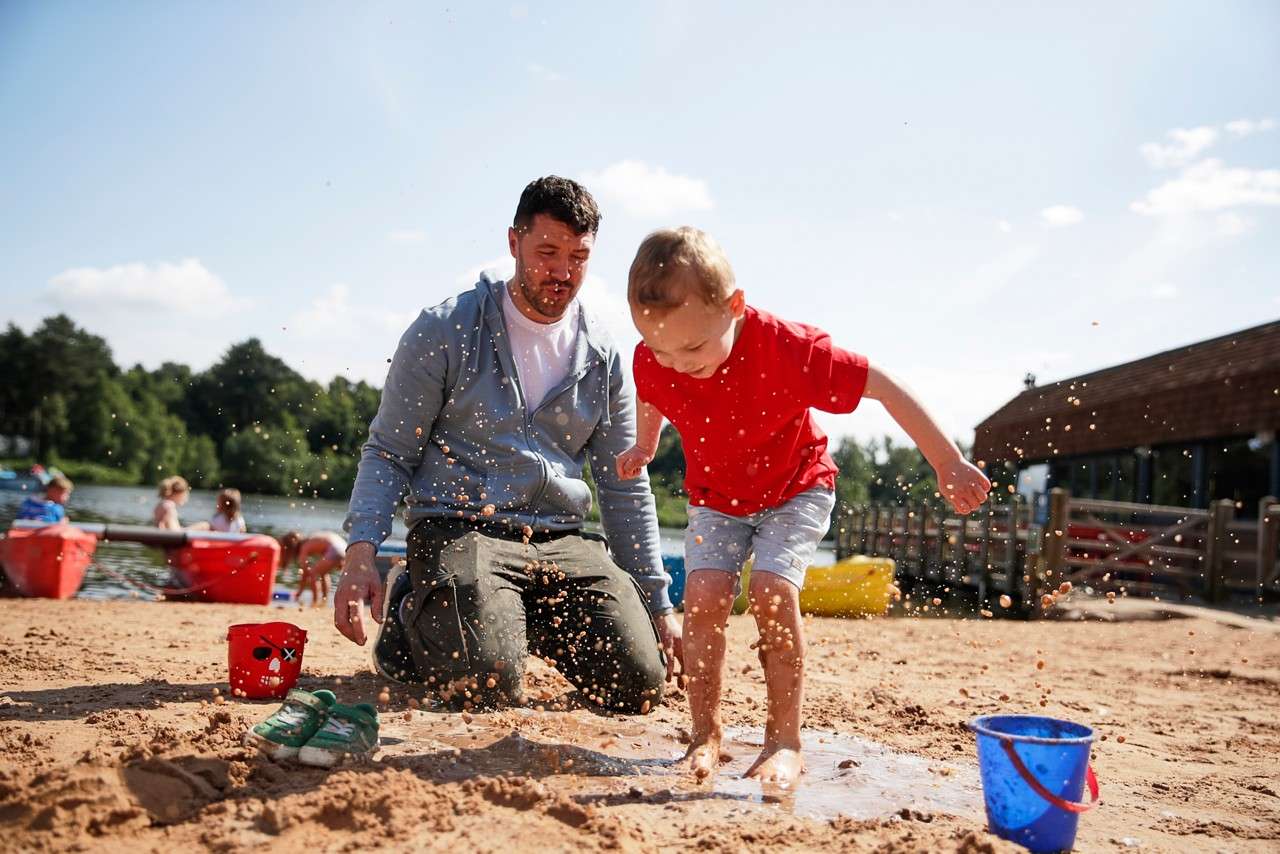 Child splashes in a sandy puddle, jumping barefoot. Adult kneels nearby watching. Around them: buckets, small shoes, kayaks, and people by a lake, with trees and buildings under a bright, clear sky.