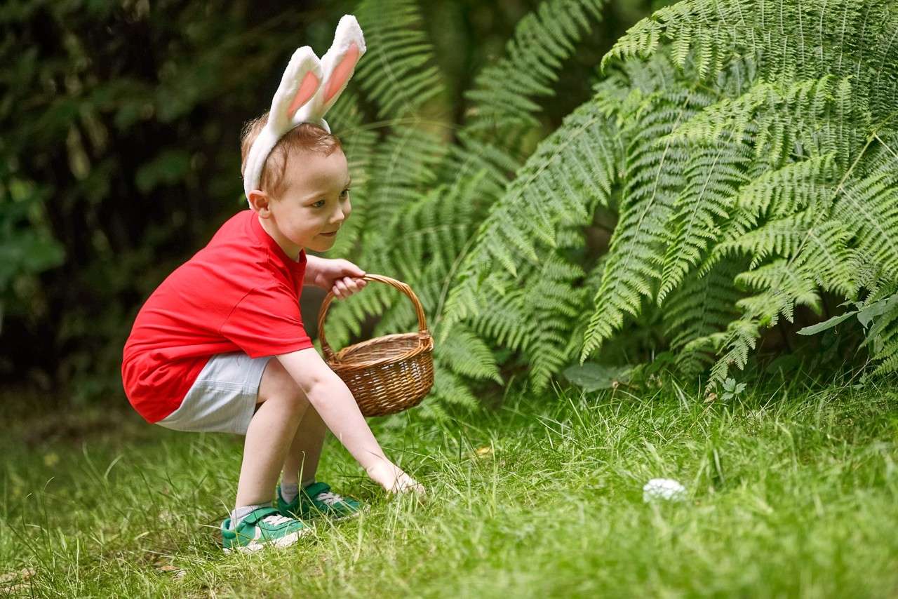 Child in a red shirt crouches, holding a wicker basket, reaching toward the grass; wearing bunny ears headband; outdoors beside large green ferns in a garden.