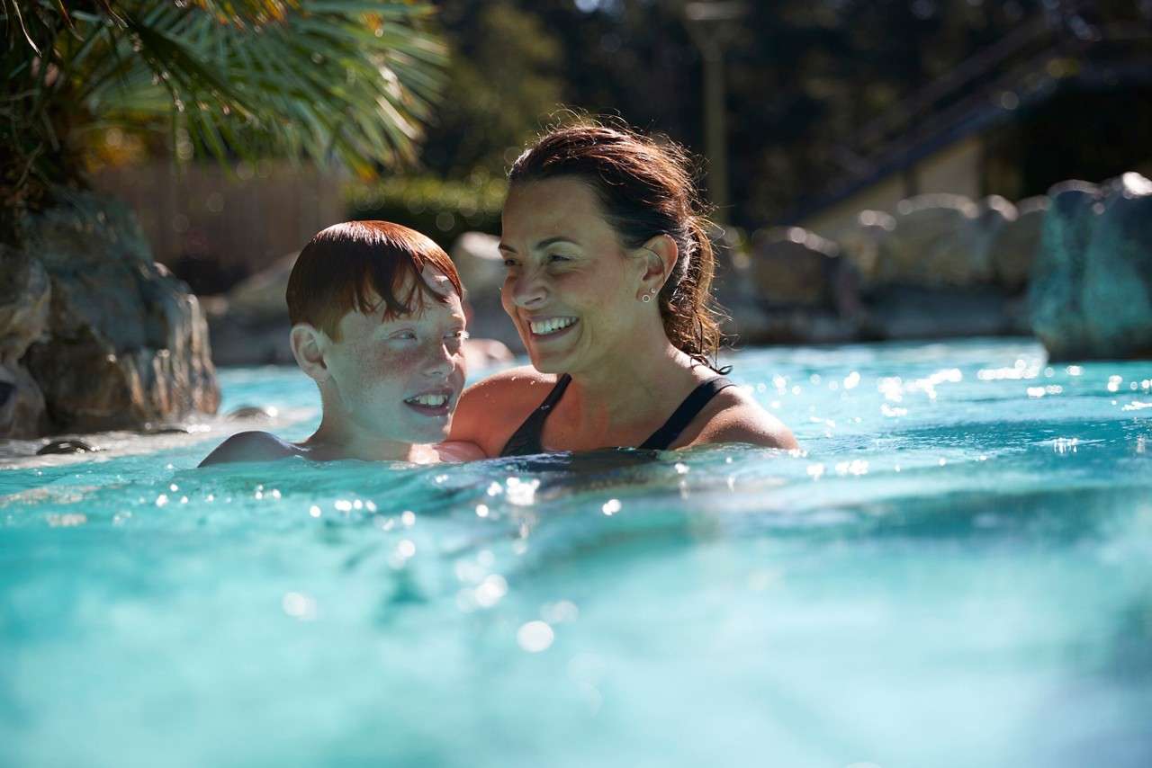 Two people smile and talk while wading in a turquoise outdoor pool, sunlight shimmering on water, with rocks and palm fronds around the edges.