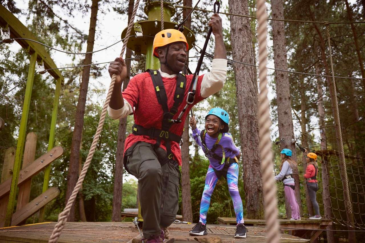 Adult and child, clipped to harnesses, balance and laugh while gripping ropes on a treetop adventure platform; two other kids wait nearby, amid tall pines, wooden platforms, and safety lines.