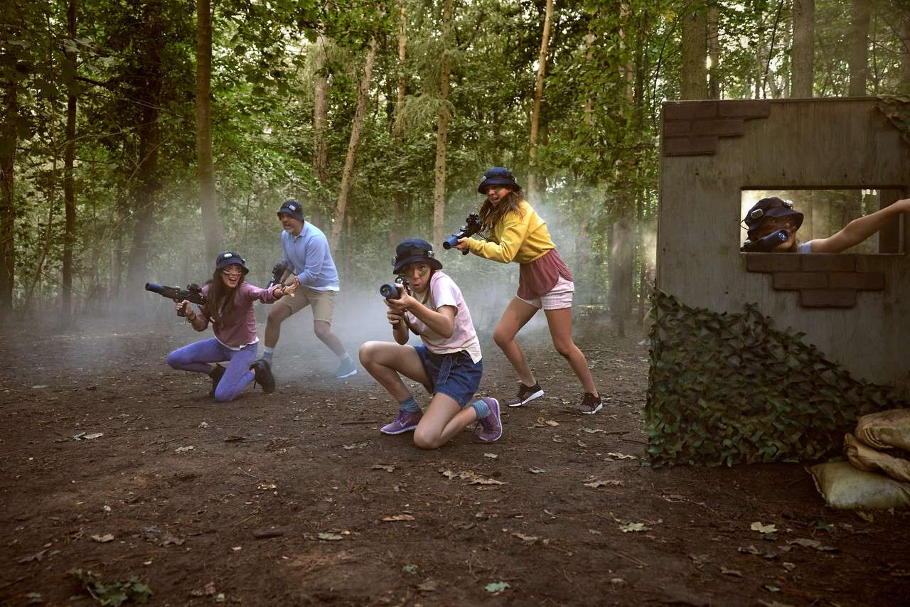 Three kids and two adults ducking and kneeling amongst the smoke in the forest holding paintball markers. One adult is partially hidden behind a wall with green camouflage leaves, pointing to a target.