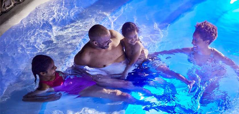 Four people wade and smile in shallow pool water; one adult holds a small child while another child lounges nearby. Blue underwater lights illuminate ripples beside a stone-edged pool.