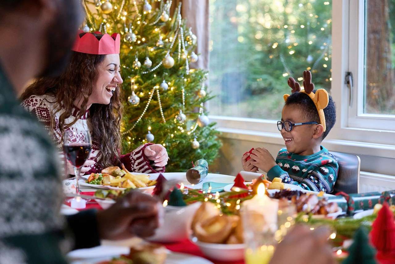 family sat around the dinner table smiling and laughing, they are all wearing christmas jumpers and paper crowns. There is a christmas tree sparkling in the background and there is lots of festive food on the table