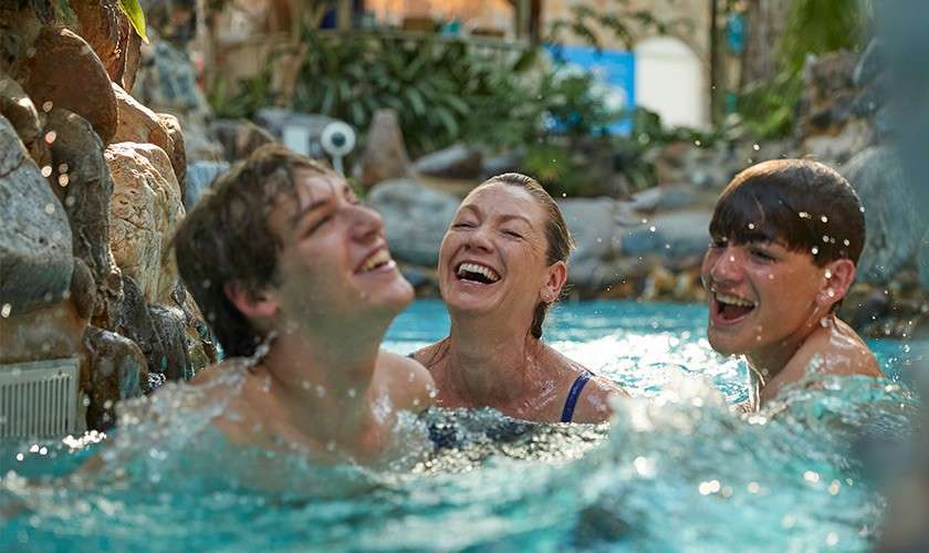 Three swimmers laugh and splash, faces upturned, as water churns around them. Set in an indoor pool with rocky edges, plants, and soft lighting, suggesting a lively water-park environment.