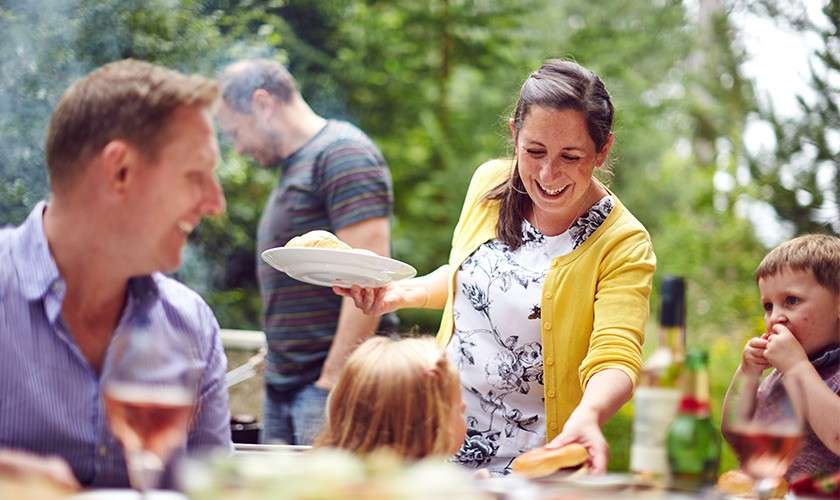 Woman in a yellow cardigan serves buns to seated children and adults, smiling. Behind her, people grill near smoky barbecue. Patio table holds drinks and food amid a green backyard.