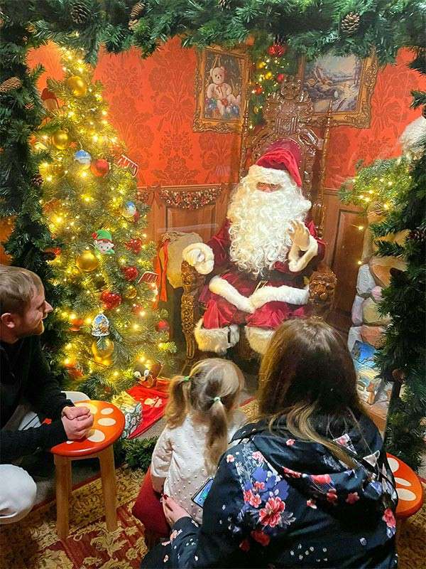 Santa Claus sits on an ornate chair, gesturing while talking to a small child. Parents kneel nearby as a lit Christmas tree, gifts, and garland fill a cozy decorated room.