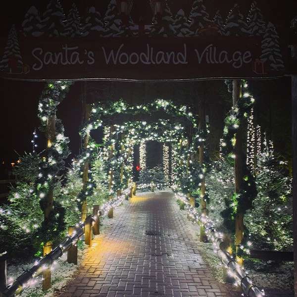 Illuminated archway glows, guiding a brick walkway; snow-dusted evergreens and white string lights decorate a nighttime path. Text: Santa's Woodland Village.