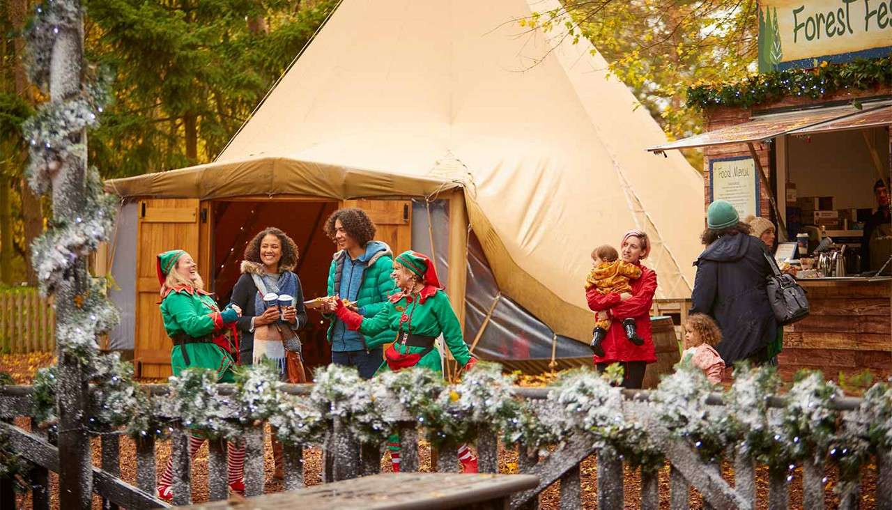 Costumed elves hand drinks to smiling visitors, chatting beside a beige teepee and wooden food stall in a forest; garlanded fence in foreground. Visible text: "Forest Fe...", "Food Menu".