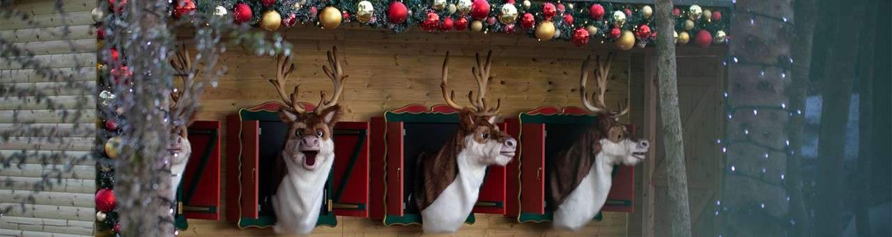Animatronic reindeer heads lean from shuttered windows, mouths open as if singing, in a wooden hut festooned with garlands and red-gold ornaments, set among trees with string lights.
