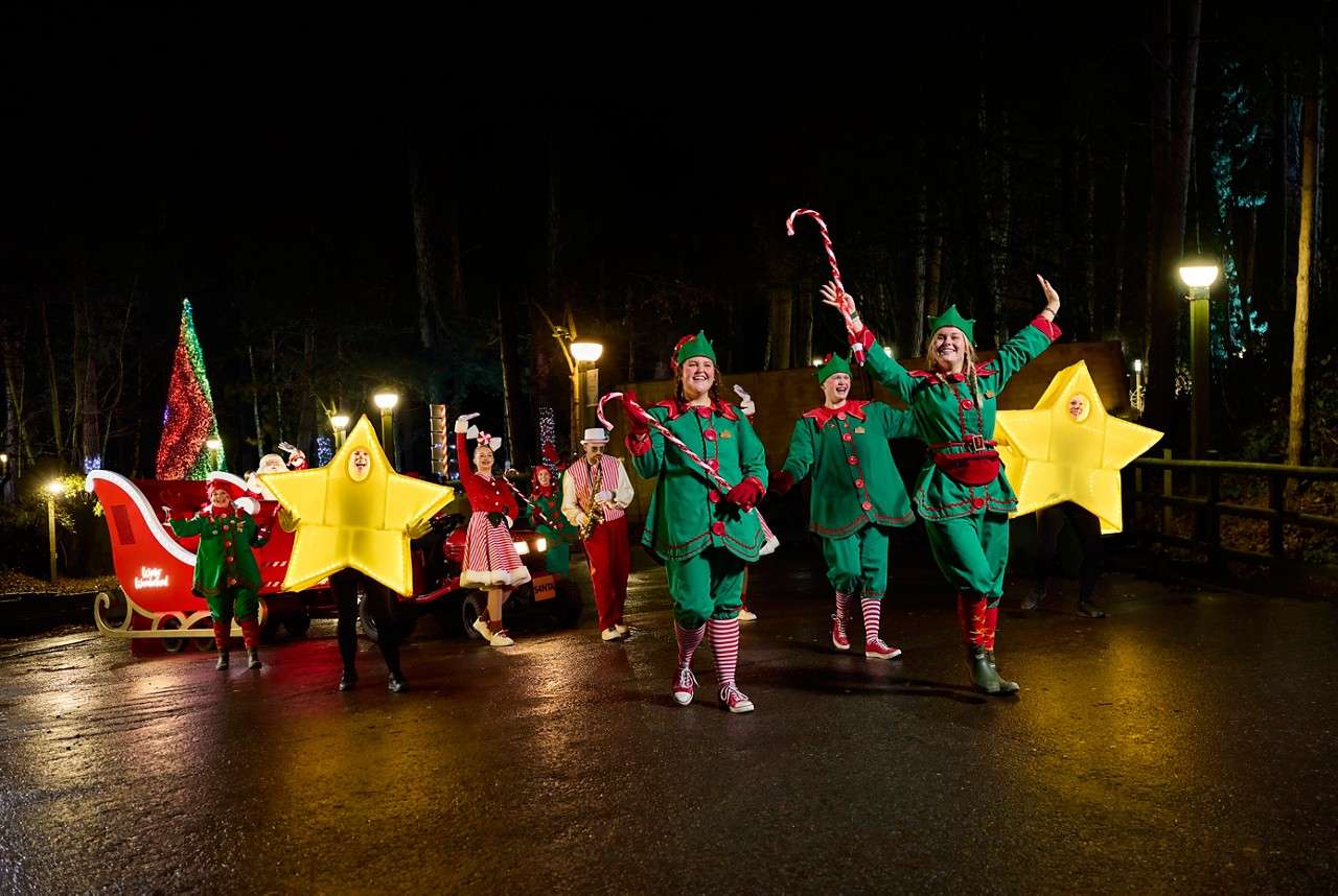 Animatronic reindeer heads lean from shuttered windows, mouths open as if singing, in a wooden hut festooned with garlands and red-gold ornaments, set among trees with string lights.
