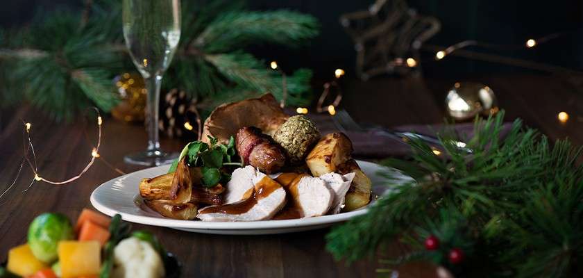 Roast turkey slices with gravy sit on a plate, accompanied by potatoes, stuffing, pigs-in-blankets, and greens, presented on a wooden table amid Christmas lights, pine branches, and a champagne flute.
