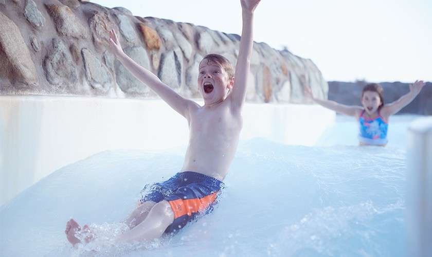 A young girl and boy sliding down the rapids with their arms in the air.
