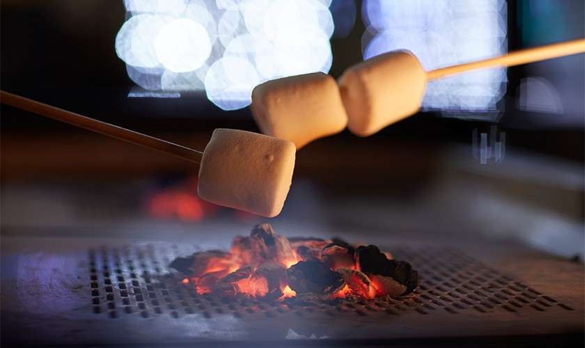 Marshmallows on wooden skewers roast over glowing charcoal on a metal grate, with blurred bokeh lights in the background, indicating an indoor or nighttime setting.