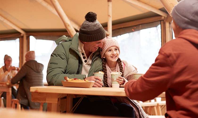 Man and girl sip hot drinks and smile at a wooden picnic table, wearing winter hats and coats, inside a rustic tent café with other patrons in background. Text: SUPERDRY.
