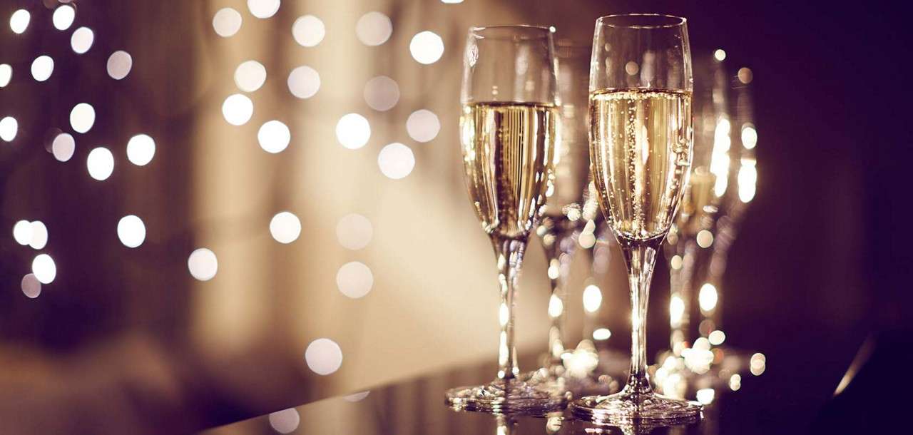 Champagne flutes stand filled with bubbling sparkling wine, catching light. They rest on a glossy table while soft, out-of-focus string lights glow in the background, creating a warm, celebratory atmosphere.