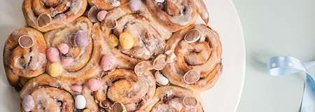 Iced cinnamon rolls cluster together, sprinkled with pastel mini chocolate eggs, sitting on a white plate on a pale surface; a light blue ribbon lies nearby.