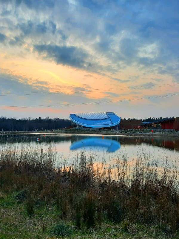Curved, blue-roofed modern building reflects on a calm lake, catching sunset light; tall grasses foreground, tree-lined shore and scattered clouds forming a tranquil, expansive setting.