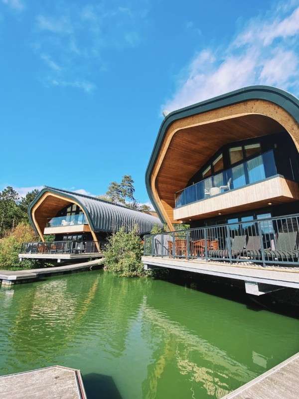 Curved-roof lakeside lodges extend over green water, their glass-fronted balconies and patio furniture facing the lagoon, set amid trees under a bright blue sky.