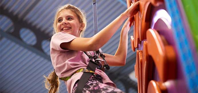 Child climber ascends, smiling, gripping a colorful indoor climbing wall; secured by harness and rope, under a high metal roof in a recreation center.