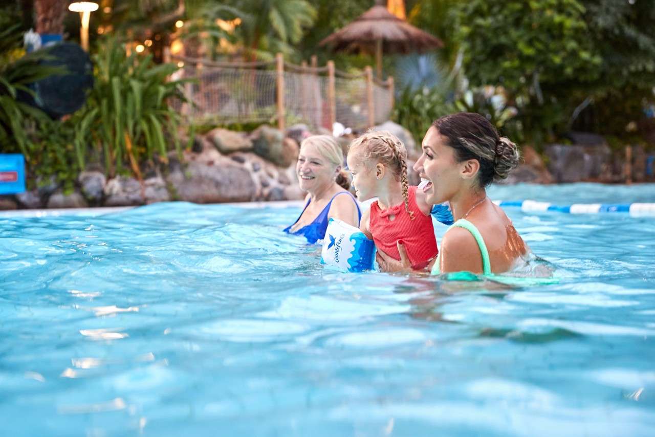 Woman holding a small child wades, smiling, beside another adult in shallow pool water; they face left. Surroundings include tropical plants, rocks, rope fence, thatched hut, and lane dividers.