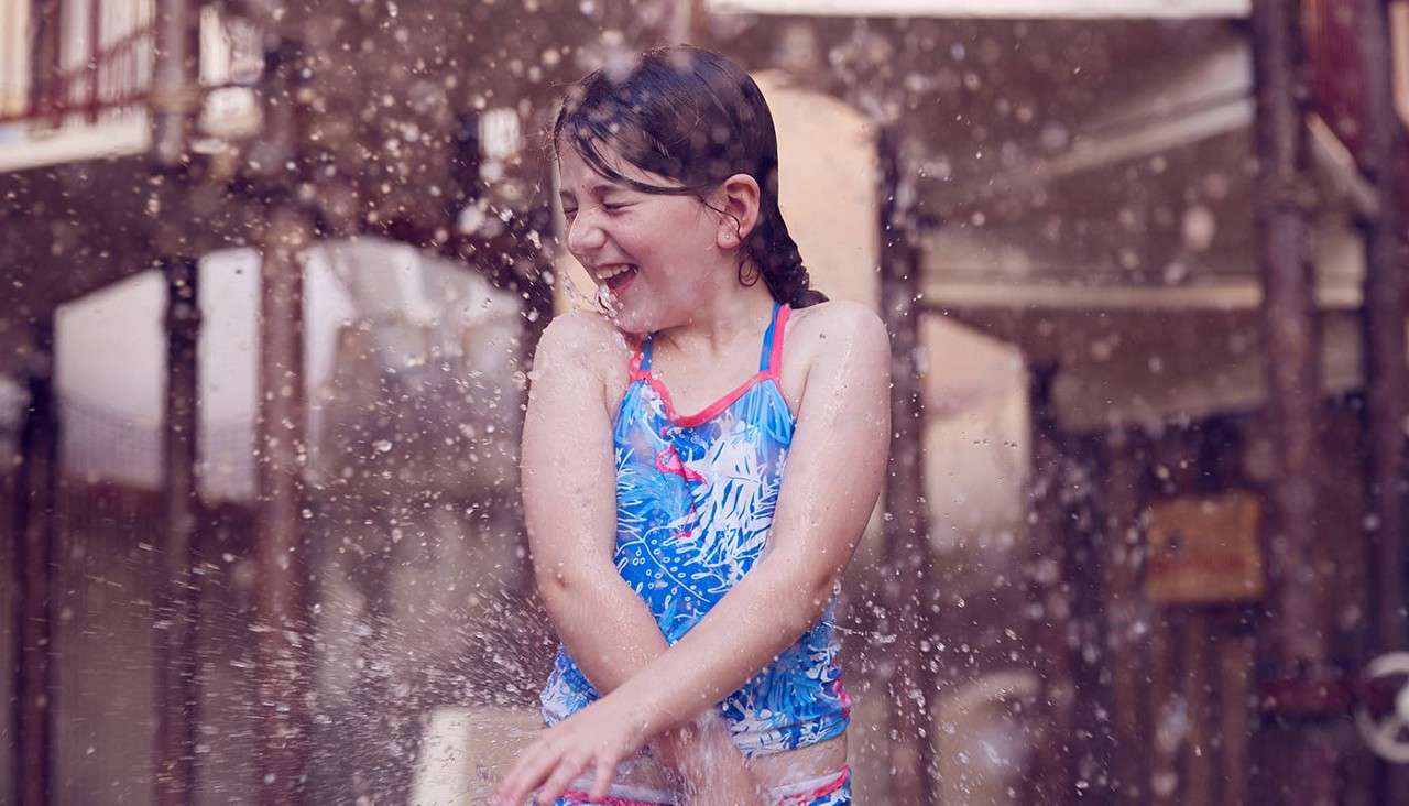 A child in a blue swimsuit laughs and braces as water splashes over their body, standing at a splash pad or water park with blurred wooden structures in the background.
