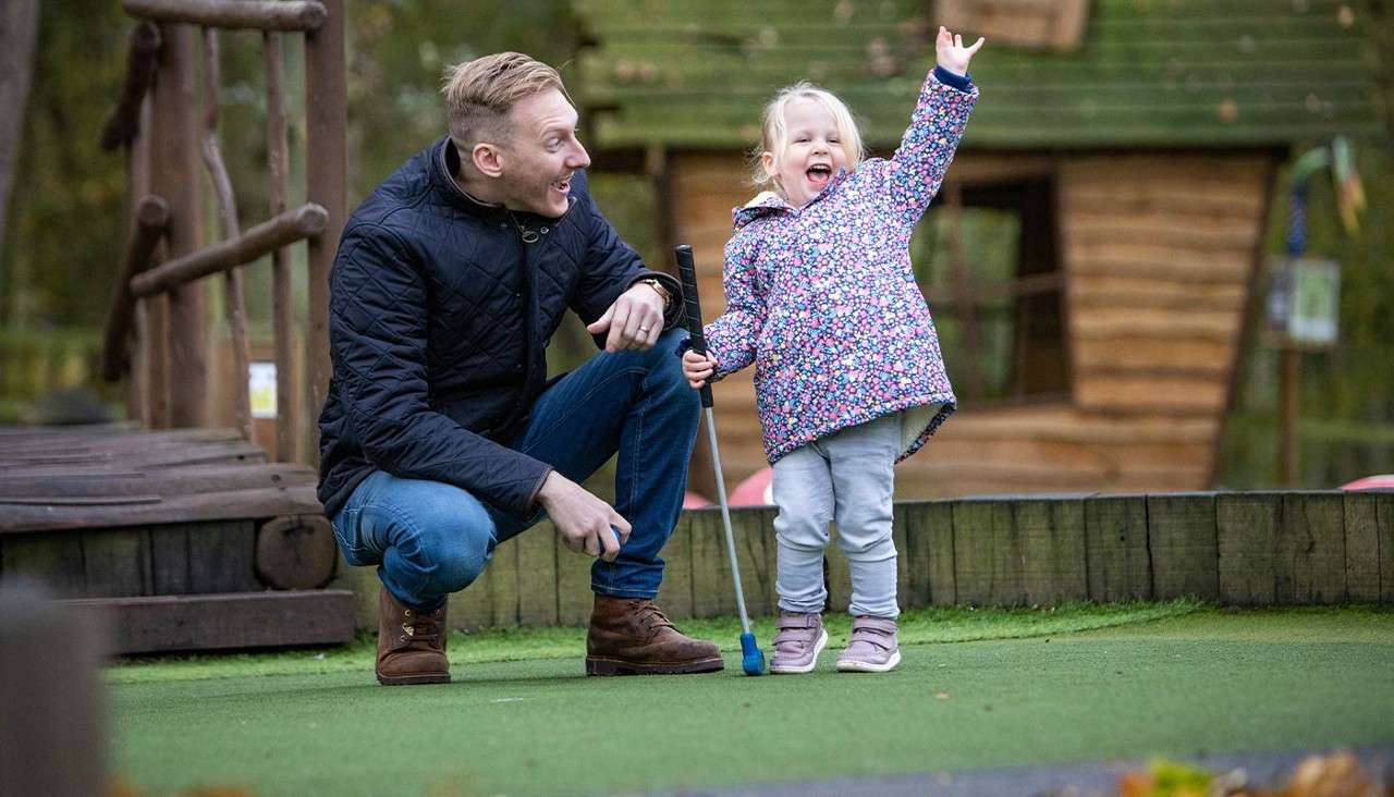 Child with mini-golf putter cheers with arms raised; man crouches beside, smiling encouragement; outdoors on a putting green near a wooden bridge and rustic playhouse amid trees.