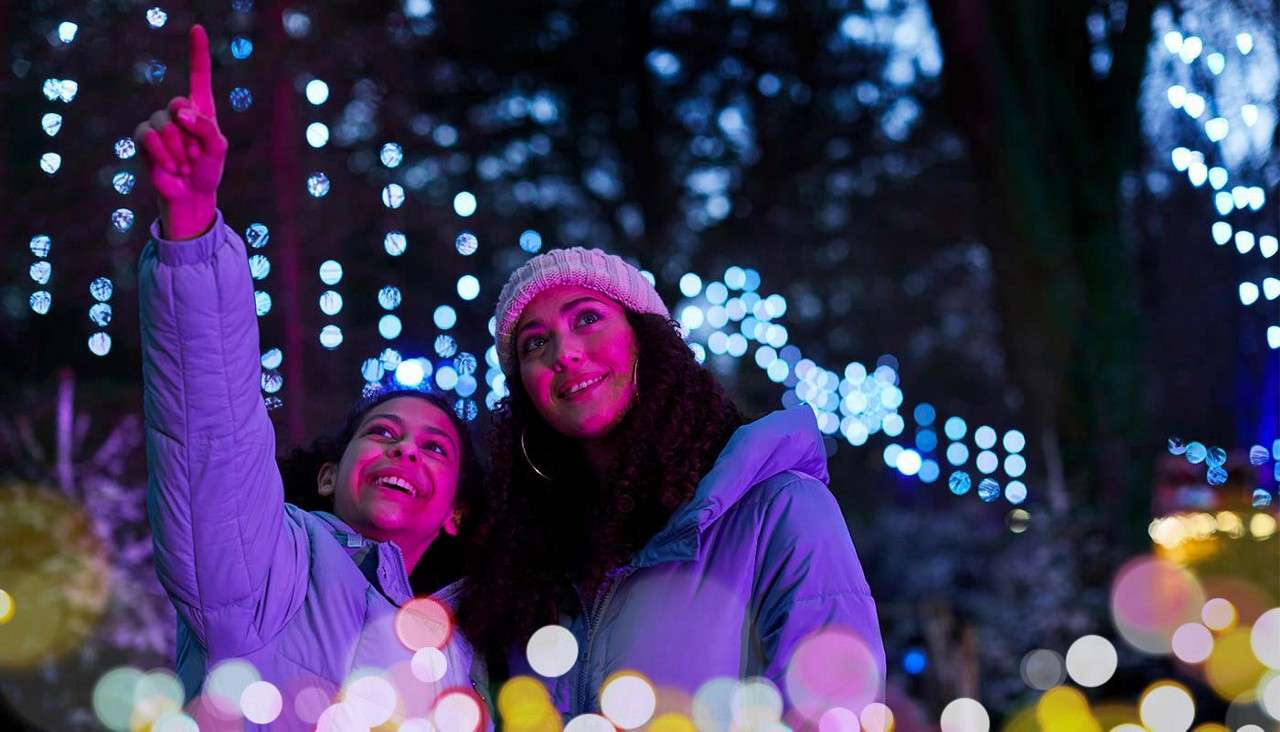 Two bundled people smile as one points upward, gazing at illuminated displays, amid a nighttime outdoor setting with blue string lights, trees, and colorful bokeh glows in the foreground.