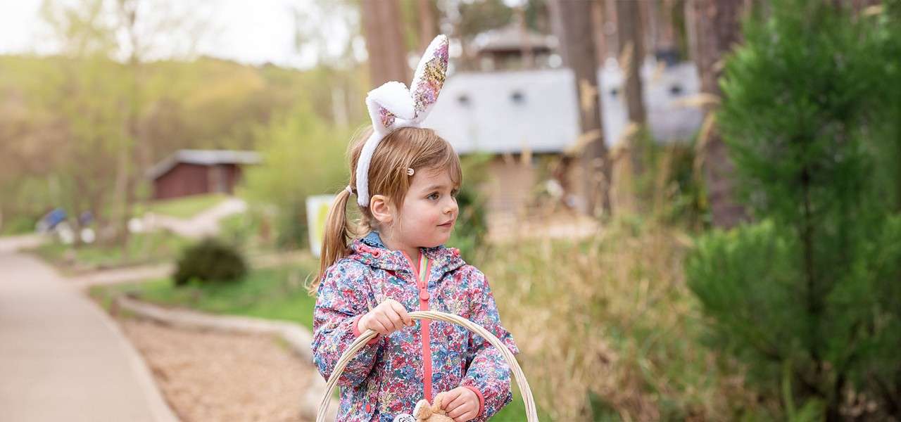 Child wearing bunny-ear headband holds a wicker basket, looking right. She stands on a park path amid trees and shrubs, dressed in a floral jacket, suggesting an Easter egg hunt.