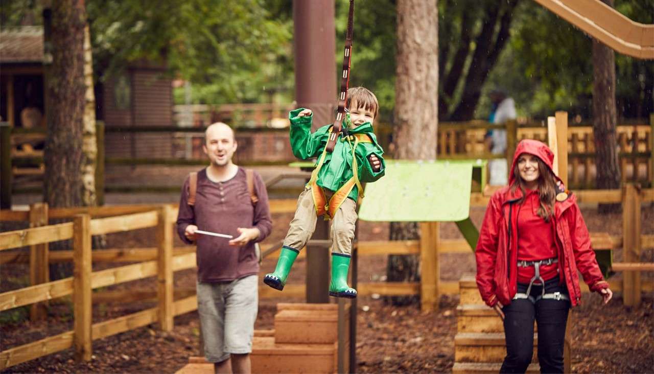 Child in green raincoat rides a harnessed zip line, feet lifted. Two adults—one casual, one in red waterproof gear—watch nearby. Wooden fences and trees indicate an outdoor adventure park.