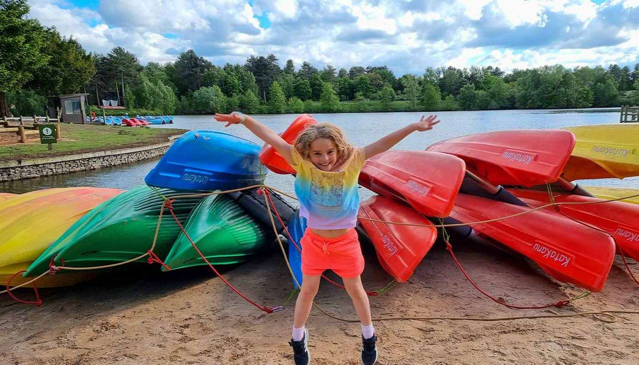 Child jumps with arms out, smiling; stacked colorful kayaks rest on a sandy lakeshore; calm lake and tree-lined park under partly cloudy sky. Text on kayaks present but unreadable.