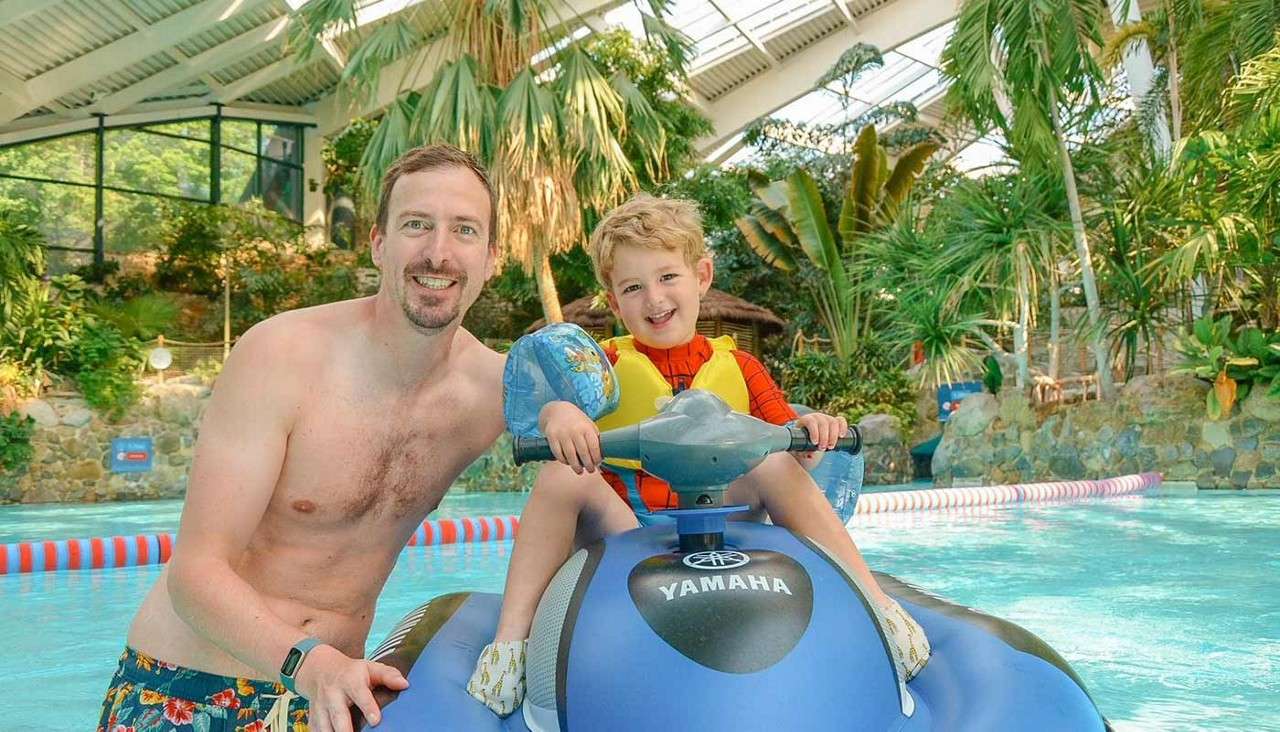 Child on blue inflatable jet ski labeled YAMAHA rides and smiles while holding handlebars; adult stands beside, both in swimwear, in an indoor tropical-themed swimming pool under a glass roof.