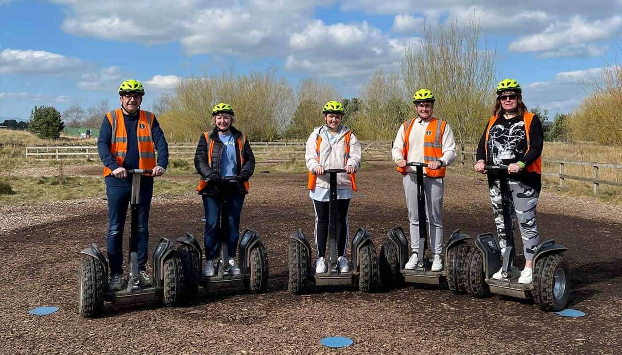 Five people on off-road Segways stand upright, facing the camera. They wear helmets and orange safety vests on a woodchip training area, with fencing, leafless trees, and blue sky.