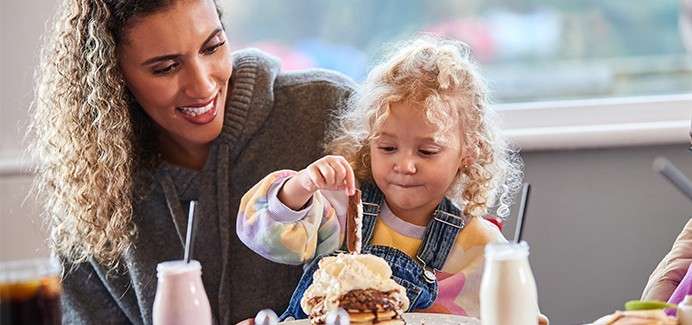 Child sprinkles toppings on pancakes with ice cream while a woman smiles beside her. At a restaurant table, milkshake bottles and drinks sit nearby, windowed background softly blurred.