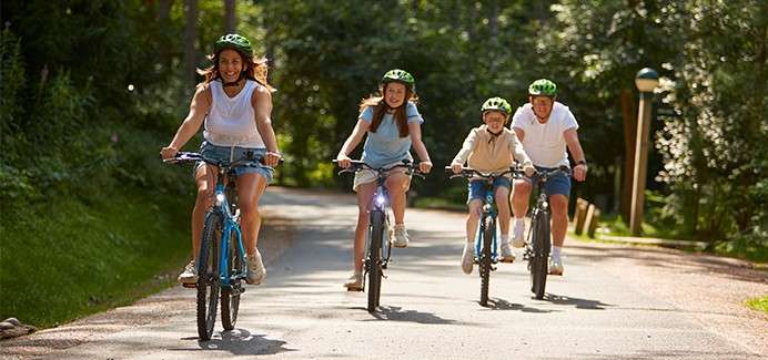 Four helmeted cyclists ride forward together, two adults and two children pedaling. They travel along a sun-dappled paved path surrounded by lush trees in a park or woodland setting.