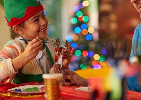 A person dressed as Santa Claus sits smiling in a warmly lit, red Christmas-themed room, surrounded by twinkling lights, ornaments, and a decorated tree in the foreground.