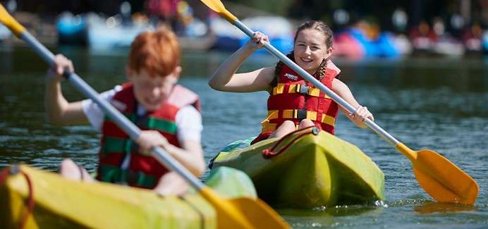 Two children paddle yellow kayaks, wearing red life jackets, smiling and focused. They stroke double-bladed paddles on a calm lake, with other colorful boats blurred in the sunny background.