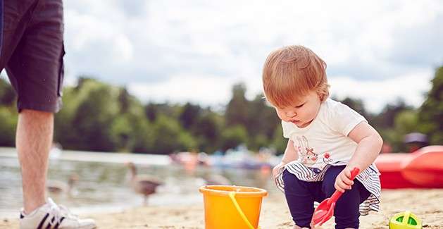Toddler crouches, scooping sand with a red shovel into an orange bucket; beside them stands an adult’s legs. Context: lakeside beach with blurred waterfowl, kayaks, trees, and cloudy sky.