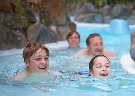 Two children swim and smile while two adults follow behind in a shallow, flowing pool, surrounded by artificial rocks and tropical greenery, suggesting a water-park lazy river setting.