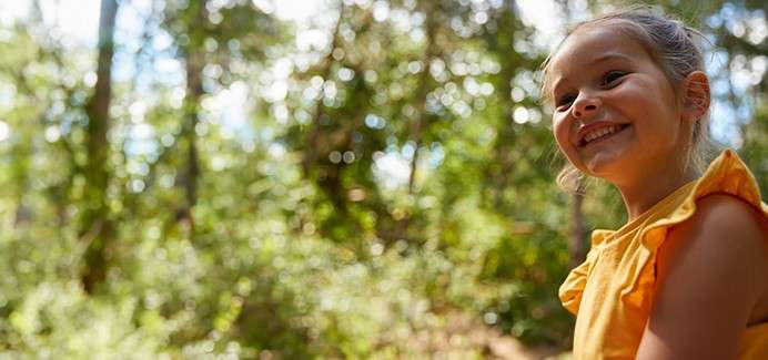 Child in a yellow sleeveless top smiles and looks right, standing in sunlit woodland with blurred trees and dappled light.