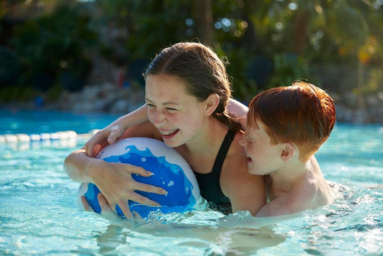 Two children embrace a blue-and-white beach ball, laughing as they play in waist-deep pool water; sunlight glints on ripples, lane rope and greenery blur in the background.