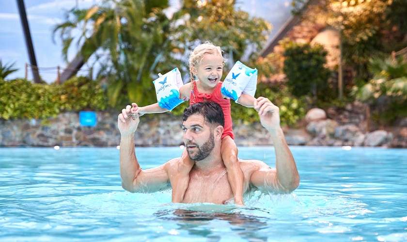 Child wearing inflatable arm floaties rides on an adult’s shoulders, both smiling and splashing gently. Context: bright outdoor pool with turquoise water, tropical plants, and stone walls in the background.
